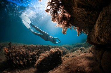 Beyaz yüzgeçli genç bir kadın güneş ışınlarıyla kayaların yakınında süzülüyor. Mavi denizde serbest dalış