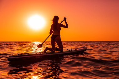 15 Temmuz 2021. Anapa, Rusya. Çekici kadın günbatımında ya da gündoğumunda sakin denizde ayağa kalk. Kırmızı yüzgeçli kadın SUP tahtası ve günbatımı.