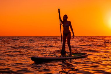 15 Temmuz 2021. Anapa, Rusya. Çekici kadın günbatımında ya da gündoğumunda sakin denizde ayağa kalk. Kırmızı yüzgeçli kadın SUP tahtası ve günbatımı.
