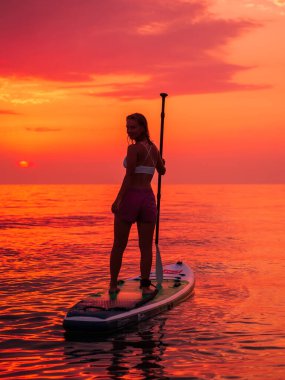 25 Haziran 2021. Anapa, Rusya. Sessiz denizde günbatımında ya da gündoğumunda kürek çeken genç bir kadın. Red Paddle yemek masasındaki kadın ve renkli günbatımı