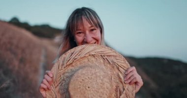 Hasır şapkalı genç bir kadın mutlu bir şekilde kameraya bakıyor günbatımında başarılı bir yaşam tarzının tadını çıkarıyor.