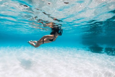Bikinili ve dalış maskeli bir kadın tropik şeffaf okyanusta kumlu zeminde suyun altında yüzüyor.