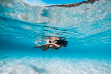 Bikinili genç bir kadın, tropikal mavi okyanusta, beyaz kum tabanlı suda yüzüyor.