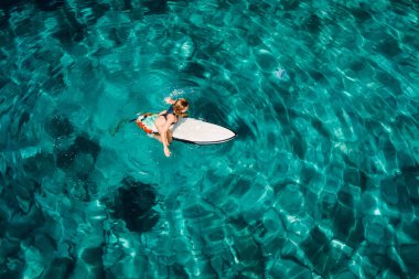 Surf girl sitting on surfboard and wait wave in turquoise ocean. Aerial view with surfer woman
