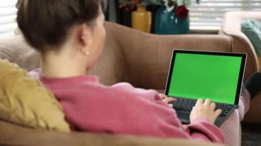 Genç bir kadın, evinde kanepede dinlenen ve dizüstü bilgisayar kullanan bir kız. Evde dizüstü bilgisayardan film izleyen bir kızın arka görüntüsü. Modern laptopta 4K ile çalışan genç bir kadının omuz üstü görüntüsü üzerinde yeşil ekran.