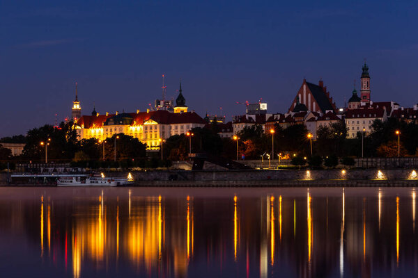 Иллюминированный пейзаж Старого города Варшавы с Королевским замком, собором и средневековыми зданиями, ночной вид с отражениями в спокойной реке Висла, Польша.