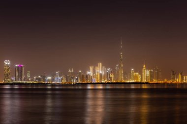 Dubai Business Bay geceleri renkli aydınlık binalar ve sakin Dubai Creek suyu ile gökyüzü çizgisi.