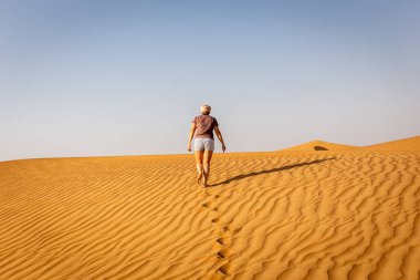 Şortlu ve tişörtlü genç beyaz kadın Arap çölüne doğru kum tepeciklerinde tek başına yürüyor, kumdaki ayak izleri ve dalgalanmalar, Birleşik Arap Emirlikleri..