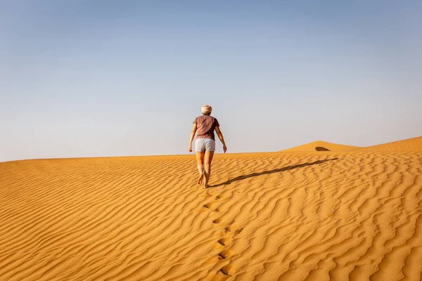 Young caucasian woman in shorts and t-shirt walking alone on a sand dune towards the arabian desert, footprints and ripples in the sand, United Arab Emirates.