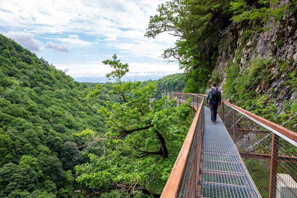 Male traveler in Okatse Canyon in Georgia, walking on hanging metal pedestrian pathway trail above deep precipice, lush vegetation and forests.