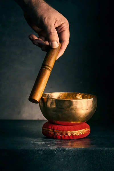 hand with a hammer and a bronze Tibetan bowl on a red pad on a dark background