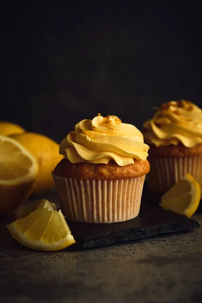 stock image two lemon cakes next to slices of yellow lemon on a dark background