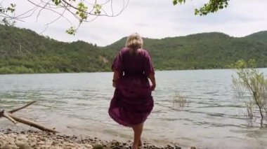 Gölün ya da nehrin kıyısında elbise giymiş genç bir kız 4K 60fps 'de filme alındı.