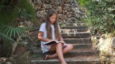 a girl with a book is sitting on the wooden steps in the garden