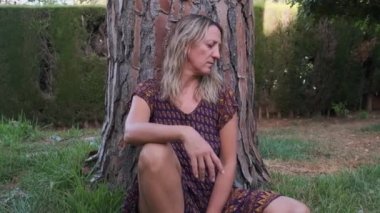 a woman in a dress is sitting on the ground near a large pine tree