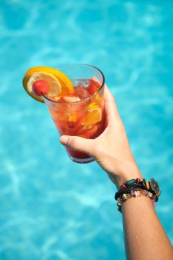 Kadın eli ve bir bardak meyve kokteyli. Havuzun arka planında portakal ve çilek var.