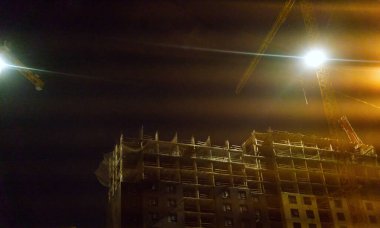 Yüksek bir binanın inşaatı. İyi geceler. İnşaat vinçleri ve evin kendisi görülebilir durumda. Işıklar parlıyor.