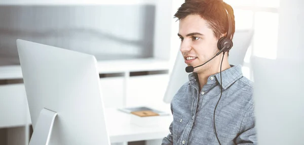 Zwyczajnie ubrany młody człowiek za pomocą słuchawki i komputera podczas rozmowy z klientami online w słonecznym biurze. Call center, koncepcja biznesowa Zdjęcie Stockowe