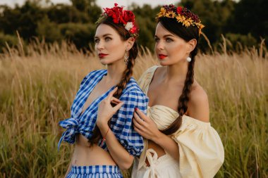 Beyaz ve mavi elbiseli genç kızlar doğanın arka planına karşı poz verirler ve ellerinde bir sepet çiçek tutarlar..