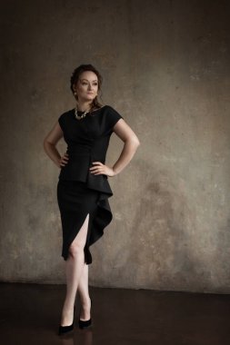 İç mekan fotoğraf stüdyosunda poz veren güzel bir genç kız. Kız klasik kesim siyah bir elbise giyiyor, saçı bir saç stiline bürünüyor ve makyaj yapılıyor. Kızın boynunda altın renkli bir zincir ve kulaklarında küpeler var..