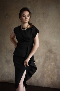 İç mekan fotoğraf stüdyosunda poz veren güzel bir genç kız. Kız klasik kesim siyah bir elbise giyiyor, saçı bir saç stiline bürünüyor ve makyaj yapılıyor. Kızın boynunda altın renkli bir zincir ve kulaklarında küpeler var..