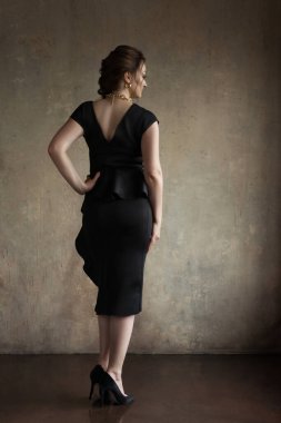 İç mekan fotoğraf stüdyosunda poz veren güzel bir genç kız. Kız klasik kesim siyah bir elbise giyiyor, saçı bir saç stiline bürünüyor ve makyaj yapılıyor. Kızın boynunda altın renkli bir zincir ve kulaklarında küpeler var..