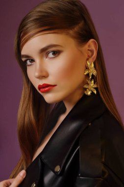 A young beautiful girl posing in a black leather jacket and gold jewelry on a background in a photo studio.