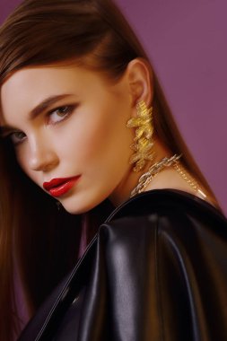 A young beautiful girl posing in a black leather jacket and gold jewelry on a background in a photo studio.