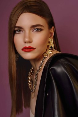 A young beautiful girl posing in a black leather jacket and gold jewelry on a background in a photo studio.