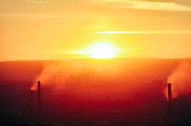 Gün batımında açık turuncu gökyüzünde elektrik santrali siluetinin bacalarından çıkan siyah zararlı duman.