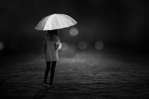A black and white portrait of a young girl walking with an umbrella in the rain alone on the street at night.