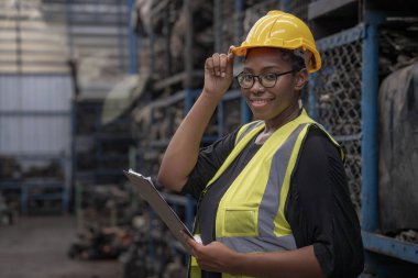 Şapka tutan ve kameraya bakan bir kadın işçi. Mühendisler veya teknisyenler depo ve fabrikalardaki otomobil parçalarını inceliyor. Afrika kökenli Amerikalı bir kadın yedek parça deposunda çizelge tutuyor..