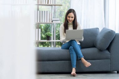 Kanepede oturan Asyalı kadın. Genç Asyalı kadın rahat koltukta dizüstü bilgisayarla çalışıyor..