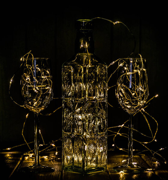 Winter celebration of Christmas, New Year and Valentine's Day. Romantic greeting. Glass bottle, wine glass and shining garland of Christmas lights