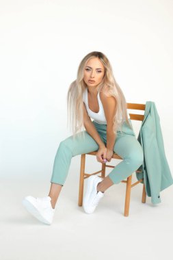 Stüdyoda, beyaz arka planda, spor ayakkabılarıyla, sandalyede oturan, nane takım elbiseli sarışın bir kızın tam boy fotoğrafı.. 