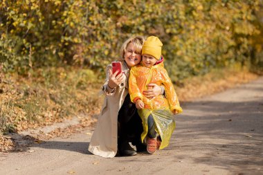 Genç bir kadın, küçük bir kızın annesi, akrabalarını aramak için akıllı telefon kullanıyor ya da sonbahar parkının ortasında selfie çekiyor.