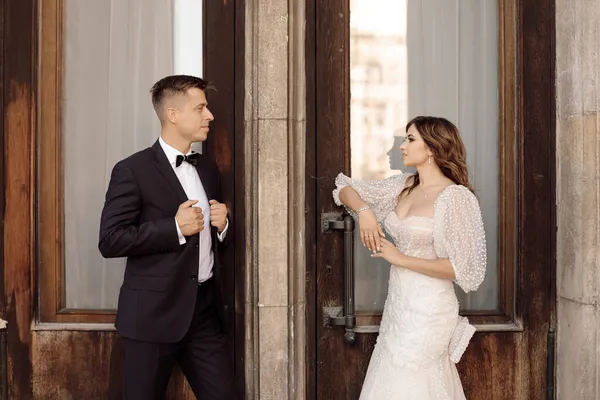 Photo of the bride and groom talking to each other and looking at each other against the background of doors. Entrance to building. Wedding dress, suit. Wedding day concept