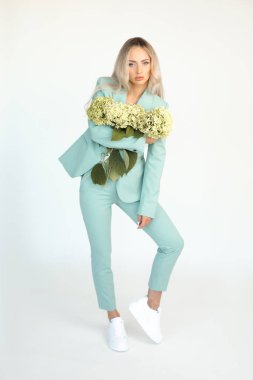 Genç, narin sarışın, uzun saçlı, nane yeşili takım elbiseli ve kolları ortanca çiçekli spor ayakkabılı, beyaz arka planda, boyunda...