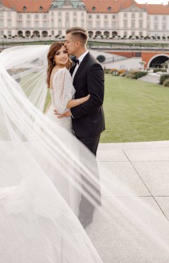 Gelinlik ve duvak takmış, kameraya bakıp gülümseyen gelin ve damat fotoğrafı. Düğün günü birlikte mutluyuz. Arka planda eski Avrupa kenti