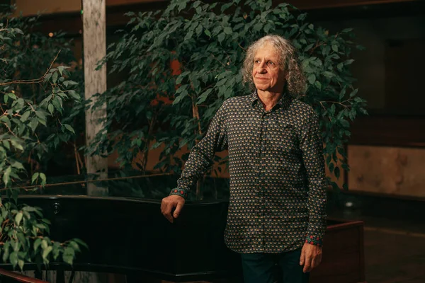 portrait of 65-year-old man at piano musical instrument in lobby of hotel among green trees gray curly hair, charismatic appearance