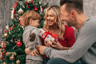Baba, anne ve oğul. Birlikte vakit geçirmek. Evde Noel ağacı, mutlu anlar. Tatile hazırlanıyorum. Birlikte mutluyuz. Hediye kutusuyla iyi eğlenceler. Kırmızı yeşil renkler. Yeni Yıl Arifesi.