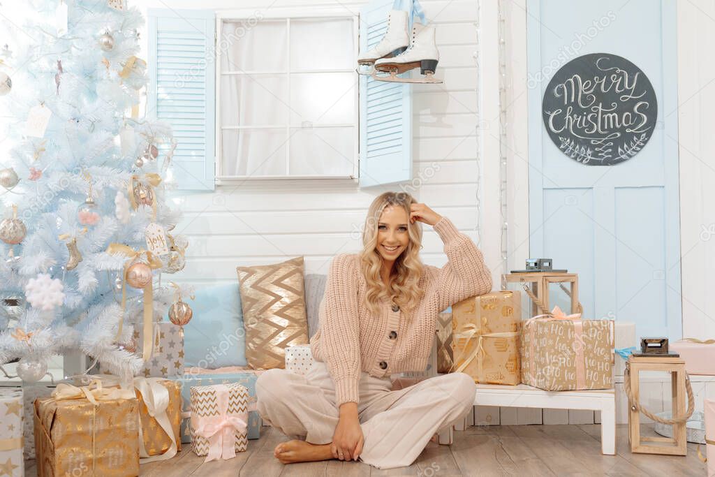Photo of young beautiful blonde sits on floor around New Year's decor and Christmas tree. In light knitted clothes and looks at camera. 