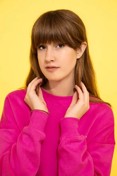 黄色の壁の長い暗い髪と前髪 茶色の目にピンクのセーターの10代の少女の肖像画は カメラを見て 手を取り合って 思春期の髪型の概念 10代の顔の皮膚のケア — ストック写真