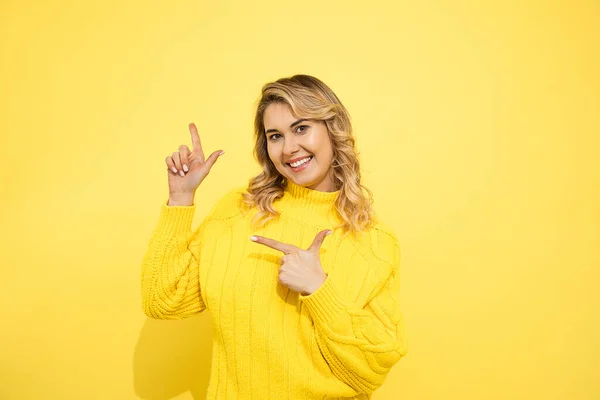 黄色の隔離された背景の上に立っている黄色のセーターを着て若いかわいい美しいブロンドの驚きの女性は 商業記号のためのスペースをコピーするために指で2本の手で指している スタジオショット — ストック写真