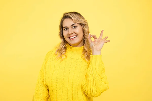 ニットセーターの若いかわいい美しいブロンドの女性は 手の指でOkに署名を示しています カメラで笑顔黄色の背景で幸せスタジオショット — ストック写真