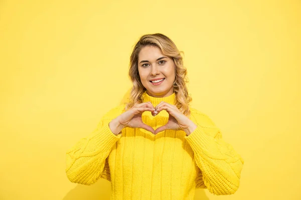黄色の壁の上にセーターを着て若いかわいい美しいブロンドの女性は ハートの形のシンボルと手で形を示す愛に笑みを浮かべて孤立した背景 デートのコンセプト スタジオショット — ストック写真
