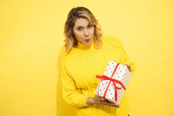 Portrait of young beautiful, successful, happy, smiling woman on yellow background holding gift box in hands, tied with red ribbon. Emotions of happiness. Interested in, enjoying holiday. Studio shot