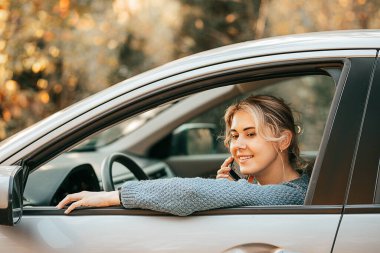 Genç, güzel sarışın bir kadının, arabayı durdururken akıllı telefonuyla konuşurken gülümsediği bir fotoğraf. Mutlu yolculuklar, rahatlama anı, aktif yaşam tarzı, trafik kurallarına uyma.