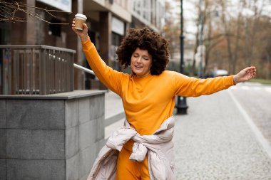 Genç, güzel beyaz kadın portresi, 40 yaşında spor kıyafetleri, sokakta tek kullanımlık kahve bardağıyla dans ediyor. Afro saç stili. Hayatın tadını çıkarıyorum, sonbahar günü. Neşeli insanlar, şehir hayatı