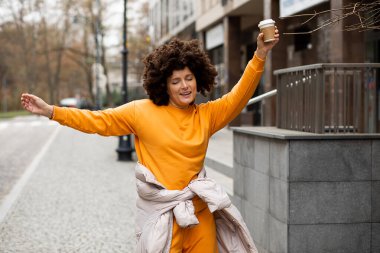 Genç, güzel beyaz kadın portresi, 40 yaşında spor kıyafetleri, sokakta tek kullanımlık kahve bardağıyla dans ediyor. Afro saç stili. Hayatın tadını çıkarıyorum, sonbahar günü. Neşeli insanlar, şehir hayatı